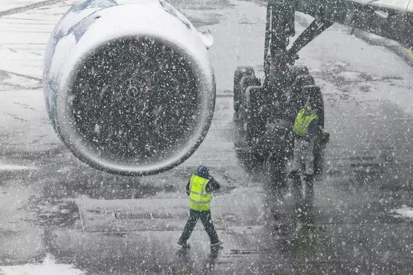 Airport staff struggling against wind and snow next to an aircraft engine