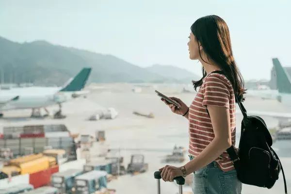 Woman standing in airport watching airplanes on runway, with phone in her hand
