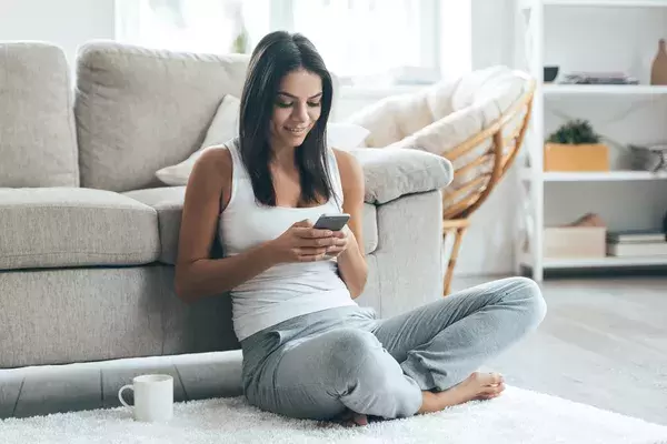 Woman sitting on the floor next to her sofa reading a message on her mobile phone