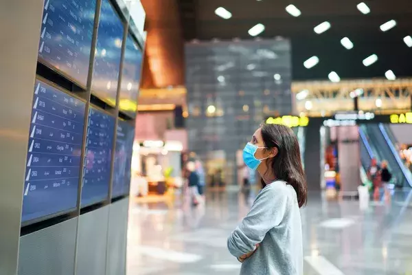 worried woman looking at departures board wearing mask in airport